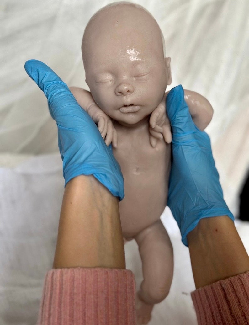 Puede incluir: Un mu&ntilde;eco de beb&eacute; de silicona realista con una tez rosa claro, ojos cerrados y boca ligeramente abierta. El mu&ntilde;eco est&aacute; siendo sostenido por manos enguantadas.