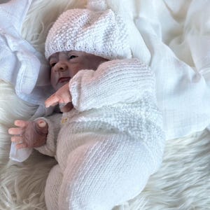 May include: A realistic newborn doll with pale skin, wearing a white knitted long-sleeved top with a single button, matching white knitted pants, and a white knitted beanie with a small pom-pom on top, resting against a soft white fabric.