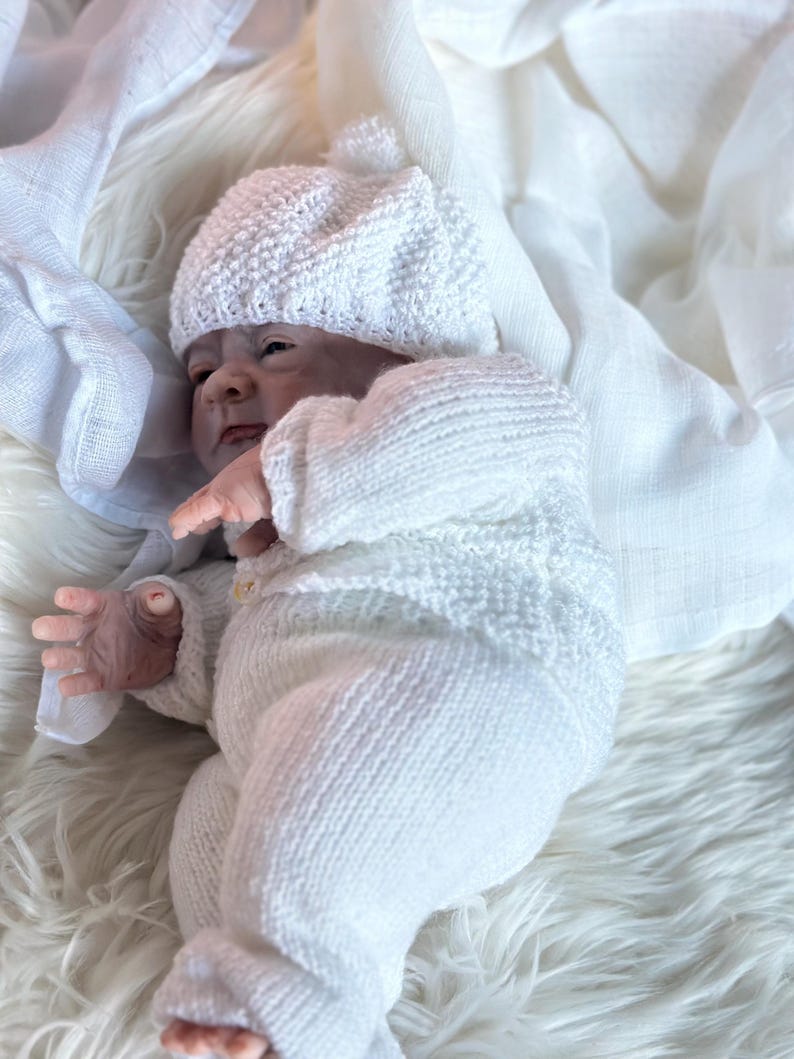 May include: A realistic newborn doll with pale skin, wearing a white knitted long-sleeved top with a single button, matching white knitted pants, and a white knitted beanie with a small pom-pom on top, resting against a soft white fabric.