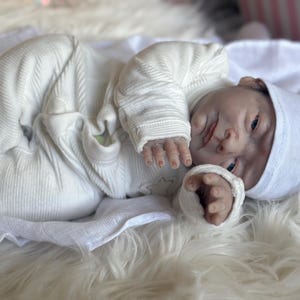 May include: A realistic newborn baby doll with pale skin, blue eyes, and detailed facial features. The doll is wearing a white long-sleeved ribbed onesie with integrated footies and a matching white knit cap, lying in a relaxed, curled position.