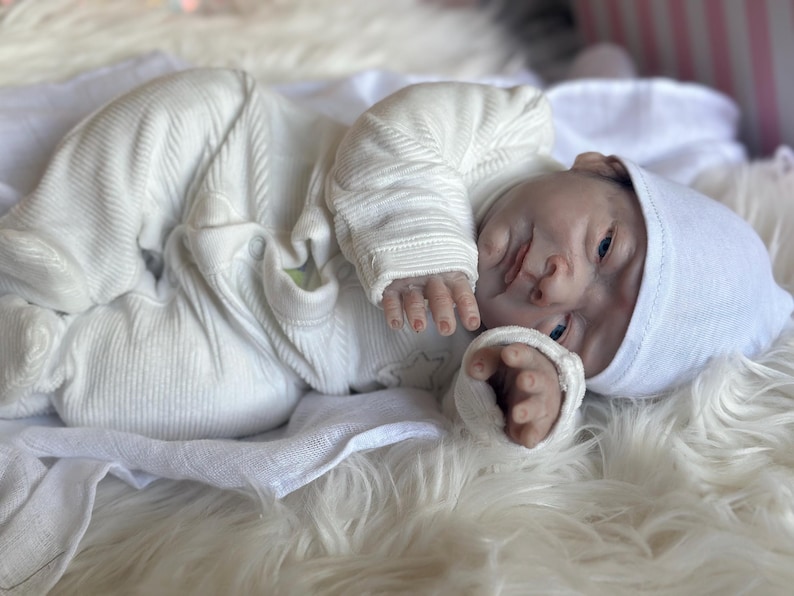 May include: A realistic newborn baby doll with pale skin, blue eyes, and detailed facial features. The doll is wearing a white long-sleeved ribbed onesie with integrated footies and a matching white knit cap, lying in a relaxed, curled position.