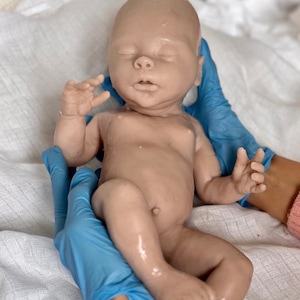 Puede incluir: Un mu&ntilde;eco beb&eacute; realista hecho de silicona, con un tono de piel marr&oacute;n claro, est&aacute; siendo sostenido por manos enguantadas. El mu&ntilde;eco tiene los ojos cerrados y la boca ligeramente abierta.