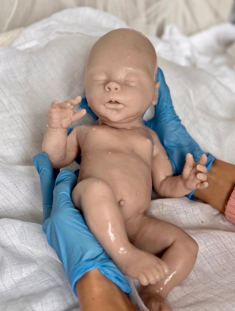 Puede incluir: Un mu&ntilde;eco beb&eacute; realista hecho de silicona, con un tono de piel marr&oacute;n claro, est&aacute; siendo sostenido por manos enguantadas. El mu&ntilde;eco tiene los ojos cerrados y la boca ligeramente abierta.