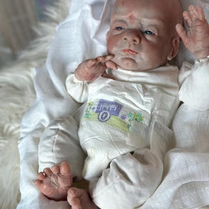 May include: A realistic newborn baby doll with pale skin, blue eyes, and a bald head. The doll is wearing a white long-sleeved shirt and a white diaper featuring a small graphic of a camper van with the word happy printed on the front.