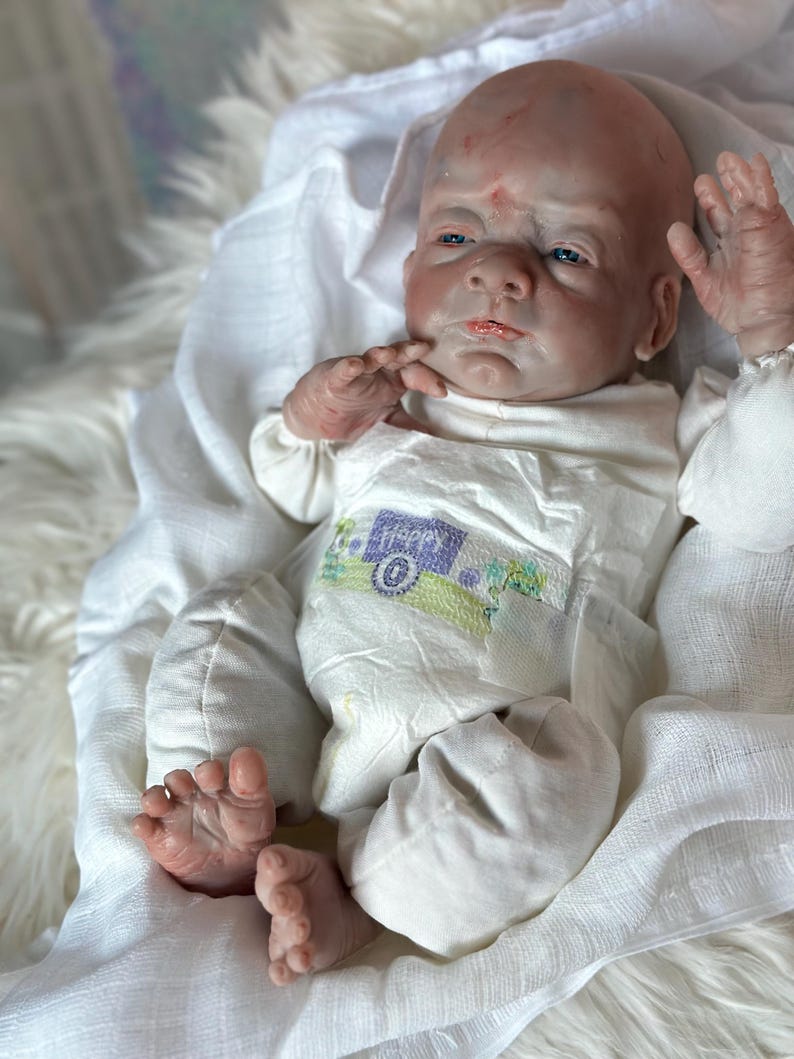 May include: A realistic newborn baby doll with pale skin, blue eyes, and a bald head. The doll is wearing a white long-sleeved shirt and a white diaper featuring a small graphic of a camper van with the word happy printed on the front.