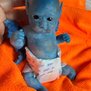 Op de afbeelding: Een blauwe fantasie babypop met puntoren en kleine blauwe vleugels. De pop draagt een luier met het woord "Happy" erop gedrukt. De pop ligt op een oranje handdoek.