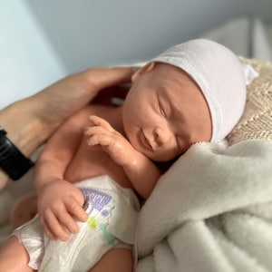 May include: A realistic newborn doll with light skin, closed eyes, and a subtle smile. The doll is wearing a plain white knit cap and a disposable diaper with a small purple and green graphic print on the front. The doll has detailed facial features.