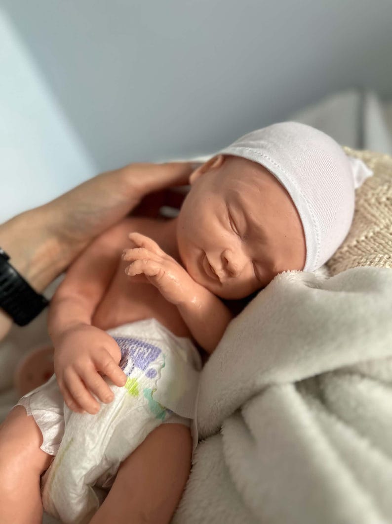 May include: A realistic newborn doll with light skin, closed eyes, and a subtle smile. The doll is wearing a plain white knit cap and a disposable diaper with a small purple and green graphic print on the front. The doll has detailed facial features.
