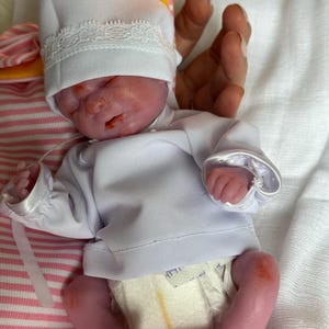 K&ouml;nnte beinhalten: Eine realistische Neugeborenen-Puppe mit rosigem Teint, bekleidet mit einem wei&szlig;en Langarmshirt, einer wei&szlig;en M&uuml;tze mit Spitzenbesatz und einer Einwegwindel. Die Puppe hat geschlossene Augen und liegt auf einem wei&szlig;en Stoff mit rosa-wei&szlig;en Streifen.