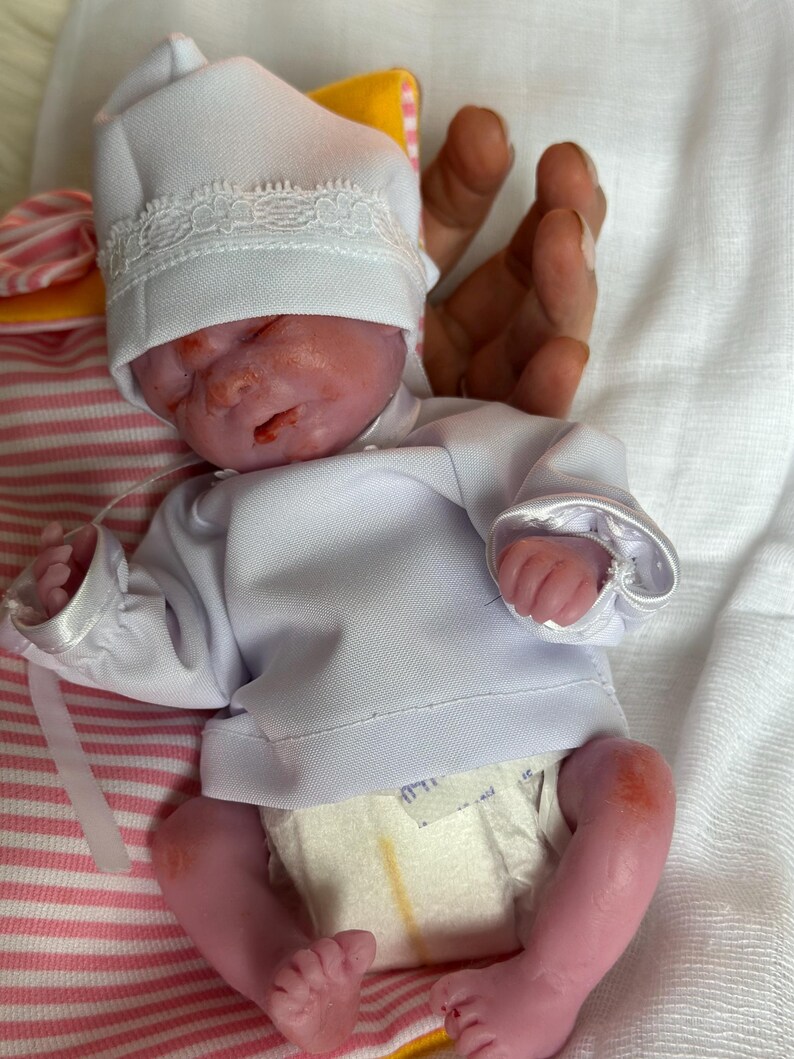 K&ouml;nnte beinhalten: Eine realistische Neugeborenen-Puppe mit rosigem Teint, bekleidet mit einem wei&szlig;en Langarmshirt, einer wei&szlig;en M&uuml;tze mit Spitzenbesatz und einer Einwegwindel. Die Puppe hat geschlossene Augen und liegt auf einem wei&szlig;en Stoff mit rosa-wei&szlig;en Streifen.