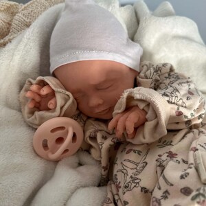 May include: A realistic baby doll with closed eyes, wearing a plain white beanie and a cream-colored long-sleeve onesie featuring a delicate floral and deer print. The doll has a light peach-colored pacifier resting near its face and hands.
