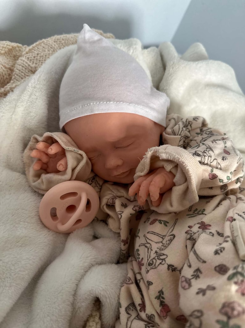 May include: A realistic baby doll with closed eyes, wearing a plain white beanie and a cream-colored long-sleeve onesie featuring a delicate floral and deer print. The doll has a light peach-colored pacifier resting near its face and hands.