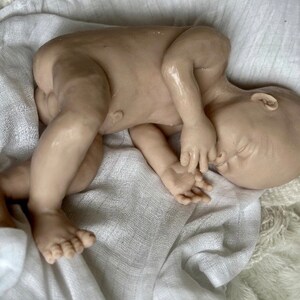 K&ouml;nnte beinhalten: Eine realistische Babypuppe mit neutralem Hautton, die auf einer wei&szlig;en, strukturierten Decke liegt. Die Puppe ist zusammengerollt, mit geschlossenen Augen und H&auml;nden in der N&auml;he des Gesichts. Die Details umfassen sichtbare Zehen und Finger.