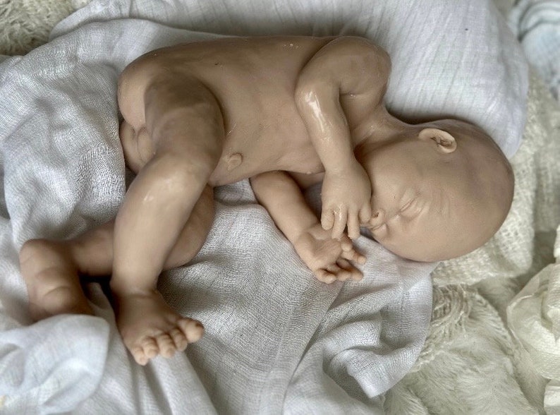 K&ouml;nnte beinhalten: Eine realistische Babypuppe mit neutralem Hautton, die auf einer wei&szlig;en, strukturierten Decke liegt. Die Puppe ist zusammengerollt, mit geschlossenen Augen und H&auml;nden in der N&auml;he des Gesichts. Die Details umfassen sichtbare Zehen und Finger.