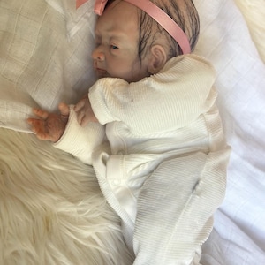May include: A realistic newborn baby doll with painted hair and a pink ribbon headband. The doll is wearing a white long-sleeved ribbed onesie with snap closures, lying on its side with one hand extended, featuring detailed skin tones and features.