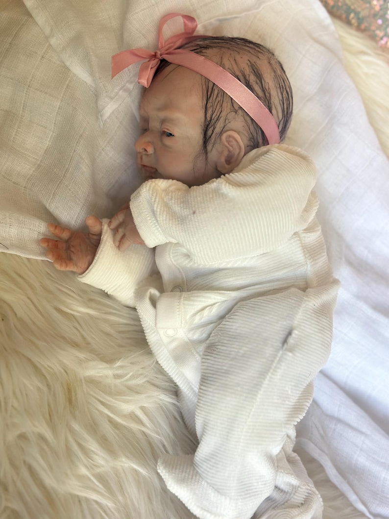 May include: A realistic newborn baby doll with painted hair and a pink ribbon headband. The doll is wearing a white long-sleeved ribbed onesie with snap closures, lying on its side with one hand extended, featuring detailed skin tones and features.