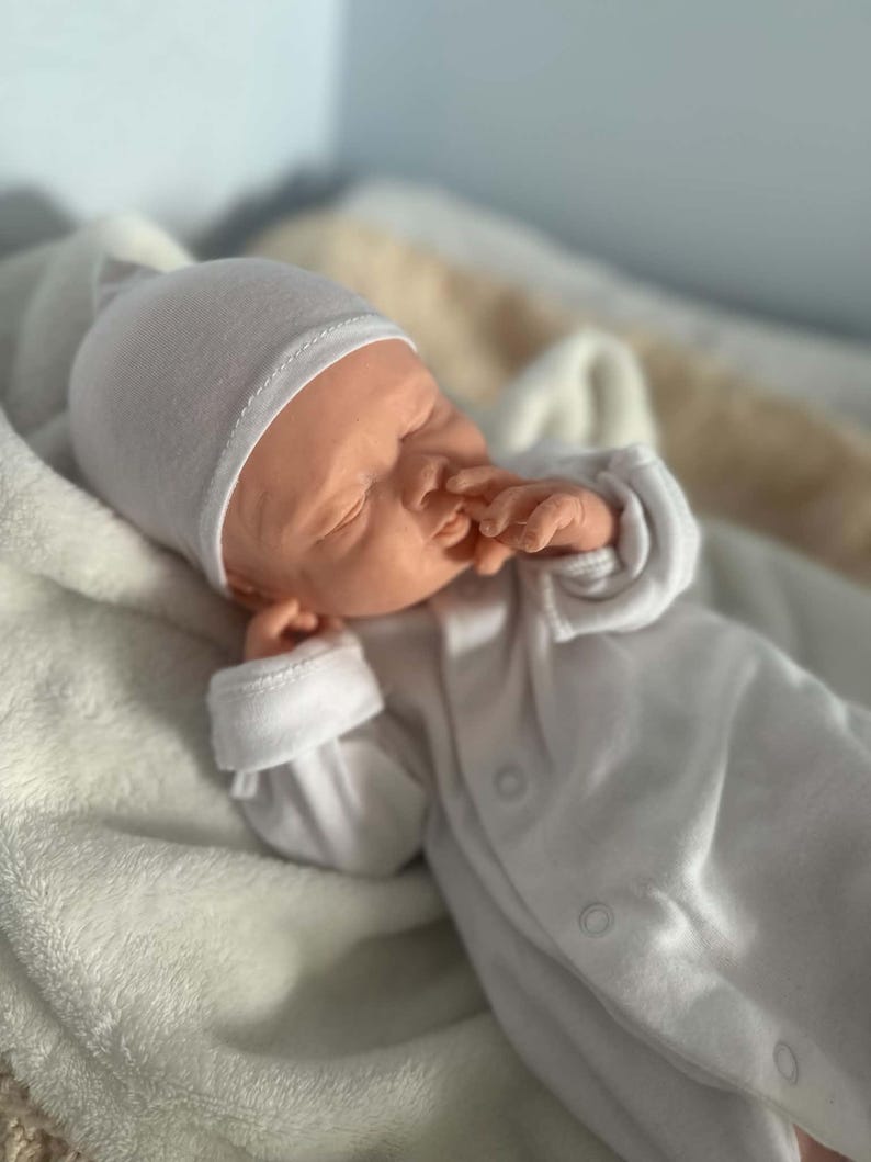 May include: A realistic newborn baby doll with light skin, closed eyes, and a small nose. The doll is wearing a plain white long-sleeved onesie with snap buttons down the front and a matching white beanie cap, resting with one hand near its face.