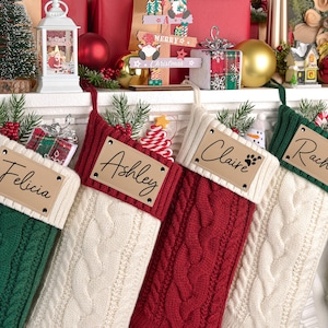 Könnte beinhalten: Vier Zopfstrick-Weihnachtsstrümpfe in Grün, Rot und Creme, jeweils mit personalisiertem Namensschild. Die Strümpfe sind mit festlichen Dekorationen gefüllt und werden auf einem weißen Kaminsims mit Weihnachtsschmuck ausgestellt.
