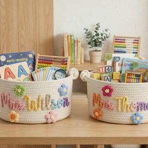May include: Two white rope baskets with colorful floral accents and embroidered names, filled with educational toys and books. One basket says "Mrs. Wilson" and the other says "Mrs. Thomas".