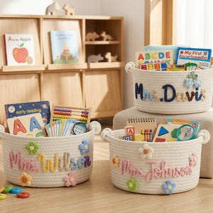 May include: Three white rope baskets with handles, filled with books and toys. Each basket has a name embroidered on it: "Mrs. Davis", "Mrs. Wilson", and "Mrs. Johnson". The baskets are decorated with colorful flower accents.