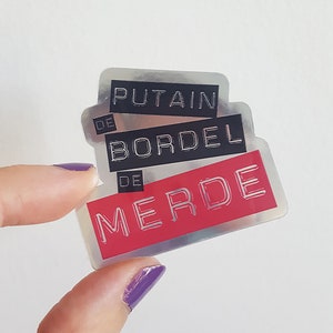 May include: A set of three stickers with French text. The top sticker reads "PUTAIN" in silver lettering on a black background. The middle sticker says "BORDEL" and the bottom sticker says "MERDE" in silver on a red background.