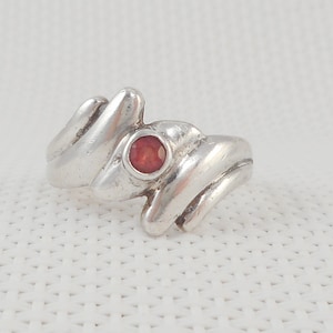 May include: A silver ring with a red gemstone set in the center. The ring has a unique, abstract design.