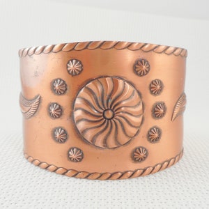 May include: A copper cuff bracelet with a swirling design in the center and a rope-like border. The bracelet is decorated with small, raised circles around the center design.