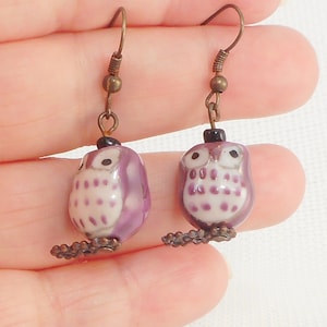 May include: Purple and white owl dangle earrings with a bronze hook and chain. The owl has black eyes and white spots on its body.