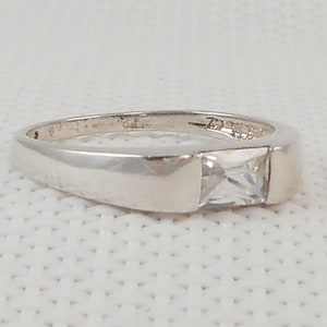 May include: A silver ring with a square, clear gemstone set in a rectangular bezel. The band is smooth and polished, with a simple, modern design. The ring is shown on a white background.