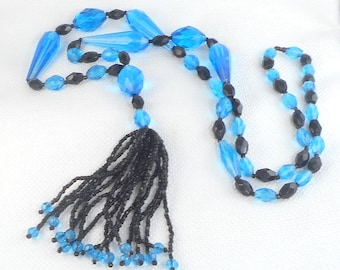 May include: A long blue and black beaded necklace with a black tassel. The necklace is made of faceted glass beads and has a long, flowing tassel.