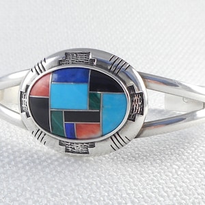 May include: A silver bracelet featuring a geometric inlay of turquoise, black, red, and green stones. The stones are set in a silver oval bezel with a decorative border.