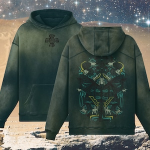 Puede incluir: Una sudadera con capucha de color verde oscuro con un aspecto descolorido y desgastado. La parte delantera presenta un pequeño diseño de pájaro negro. La parte trasera presenta un gran diseño intrincado en tonos de verde, azul y amarillo.
