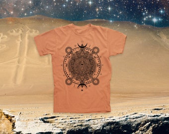 May include: A peach-colored t-shirt with a black and white graphic print of a circular design with a central eye and geometric patterns. The design is reminiscent of a mandala or a celestial map.