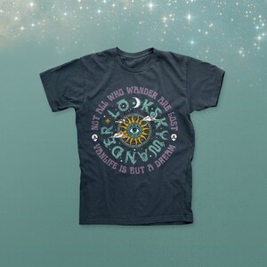 Op de afbeelding: Donkerblauw t-shirt met een cirkelvormig grafisch ontwerp. Het ontwerp toont een zon met een oog in het midden, omringd door de tekst: "Not all who wander are lost. Vanlife is but a dream."