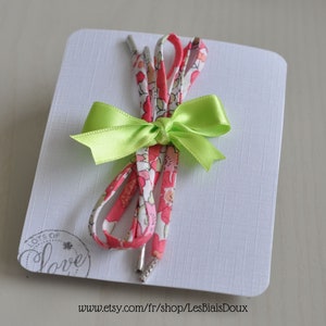 May include: A pair of shoelaces with a floral print in pink and white, tied together with a green satin bow. The shoelaces are displayed on a white card with the text "Lots of Love" and a website address.