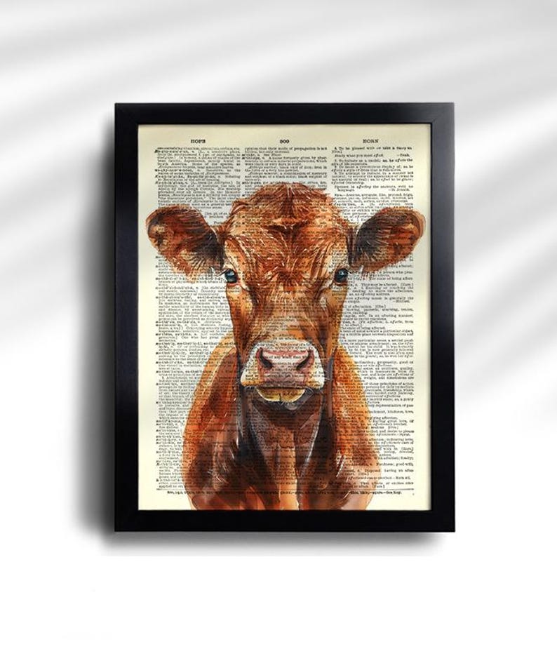 May include: A framed print featuring a watercolor painting of a brown cow. The cow is centered, with a detailed depiction of its face and fur. The background is a vintage dictionary page, adding a rustic touch to the artwork.