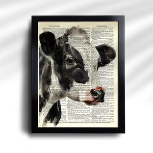 May include: Black and white cow portrait art print in a black frame. The cow's face is painted over a vintage dictionary page. The cow has a pink nose and is looking to the left. The print is a unique piece of wall decor.