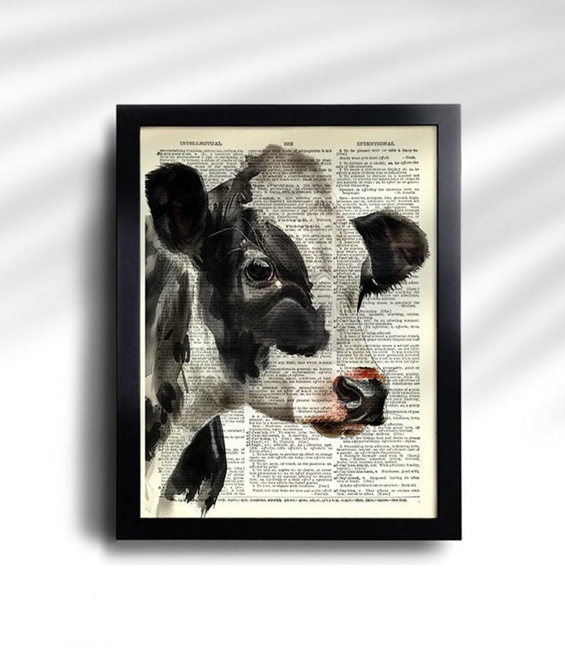 May include: Black and white cow portrait art print in a black frame. The cow's face is painted over a vintage dictionary page. The cow has a pink nose and is looking to the left. The print is a unique piece of wall decor.