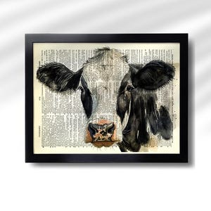 May include: Black framed art print featuring a watercolor painting of a cow's face. The cow's face is black and white, with a pink and brown nose. The background is a vintage dictionary page.