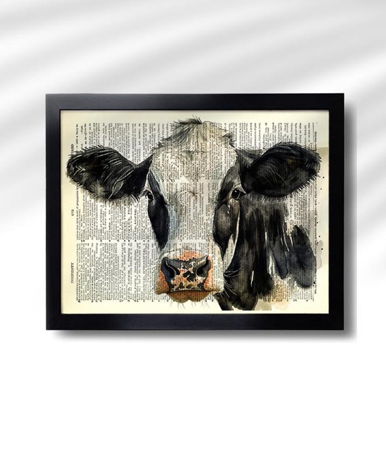 May include: Black framed art print featuring a watercolor painting of a cow's face. The cow's face is black and white, with a pink and brown nose. The background is a vintage dictionary page.