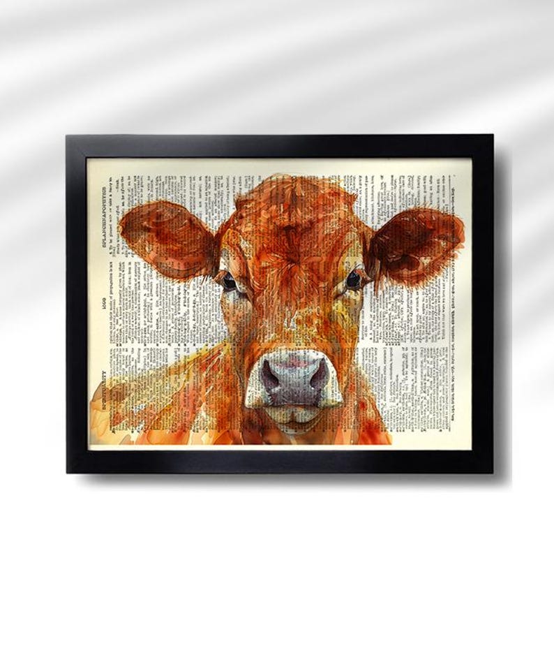May include: A framed watercolor print of a cow's head. The cow is a reddish-brown color, with large ears and a gentle expression. The background is a vintage dictionary page, and the frame is black.