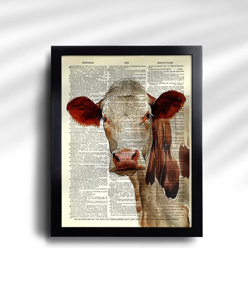 May include: Framed art print featuring a cow illustration. The cow has red ears and a brown and white face, set against a vintage dictionary page background. The black frame adds a classic touch to the artwork.