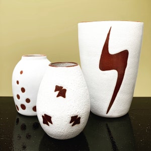 May include: Three white ceramic vases with brown abstract designs. The tallest vase has a lightning bolt design, the middle vase has a geometric design, and the smallest vase has a polka dot design.