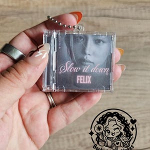 May include: A miniature clear plastic CD case keyring featuring a black and white portrait of a person. The cover art includes the text "Slow it down" in a cursive font and the name "FELIX" in bold pink capital letters below the portrait.