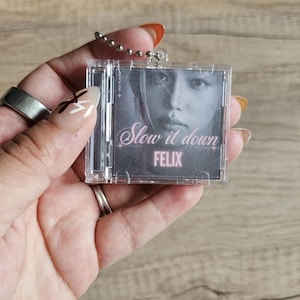 May include: A miniature clear plastic CD case keychain featuring a black and white portrait of a person. The cover art includes the text "Slow it down" in a cursive font and the name "FELIX" in bold pink capital letters below the image.