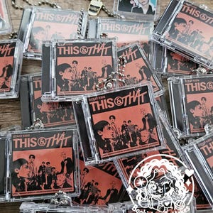May include: A collection of miniature clear plastic CD case keychains featuring the Stray Kids This & That album cover art in shades of orange and black. Each keychain includes a silver ball chain and a small charm, such as a coffee cup.