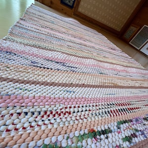 Könnte beinhalten: Ein handgefertigter, rechteckiger Flickenteppich mit Streifenmuster. Der Teppich zeigt abwechselnde Reihen in verschiedenen Farben, darunter Rosa, Weiß, Beige und Grün. Der Teppich besteht aus gewebten Stoffstreifen.
