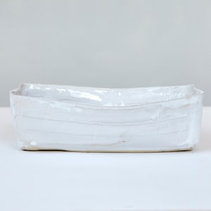 Könnte beinhalten: Eine handgefertigte, rechteckige weiße Keramikschale mit leicht gebogenem Rand. Die Schale hat eine strukturierte Oberfläche mit horizontalen Linien und ein glänzendes Finish. Das minimalistische Design eignet sich zum Servieren von Speisen oder als Dekoration.