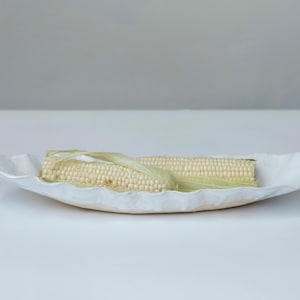 Op de afbeelding: Een witte, keramische serveerschaal met geschulpte randen bevat twee maïskolven. De maïs heeft lichtgele korrels en groene bladeren. De schaal staat op een wit oppervlak, met een neutrale achtergrond.