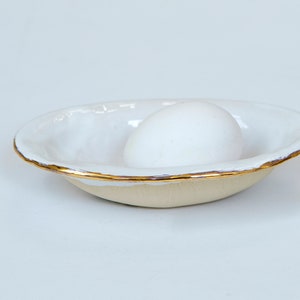 Może przedstawiać: Ręcznie robiony ceramiczny pojemnik na jajka z białym wnętrzem i beżowym zewnętrzem. Obręcz jest ozdobiona złotą metalową opaską. Białe jajko spoczywa wewnątrz miski. Miska jest przedmiotem dekoracyjnym.